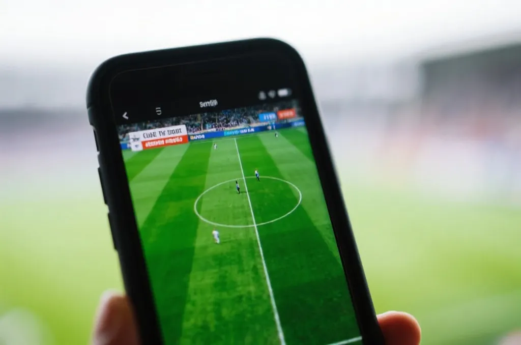 A close-up of a smartphone screen displaying a live soccer game broadcast, green grass field visible on screen